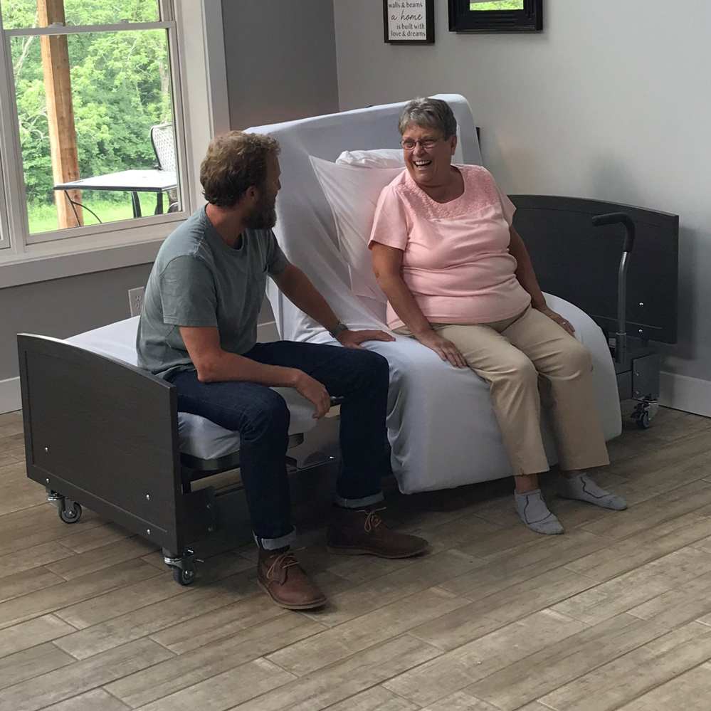 Smiling senior woman sitting upright at the edge of a Med-Mizer ActiveCare rotating bed, feet on the floor, while a caregiver kneels beside her in a home bedroom
