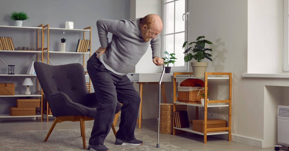 A person using their cane to stand up from a chair near a window. The person places one hand on their lower back.