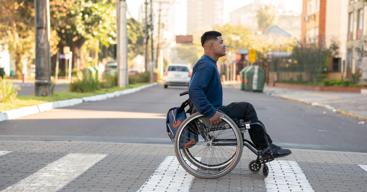 A person wearing a dark blue sweater crossing the street in a wheelchair during the daytime. A white car is nearby.