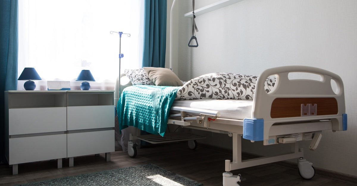 A home hospital bed installed next to a large window in the corner of a room. A blue blanket is on the hospital bed.