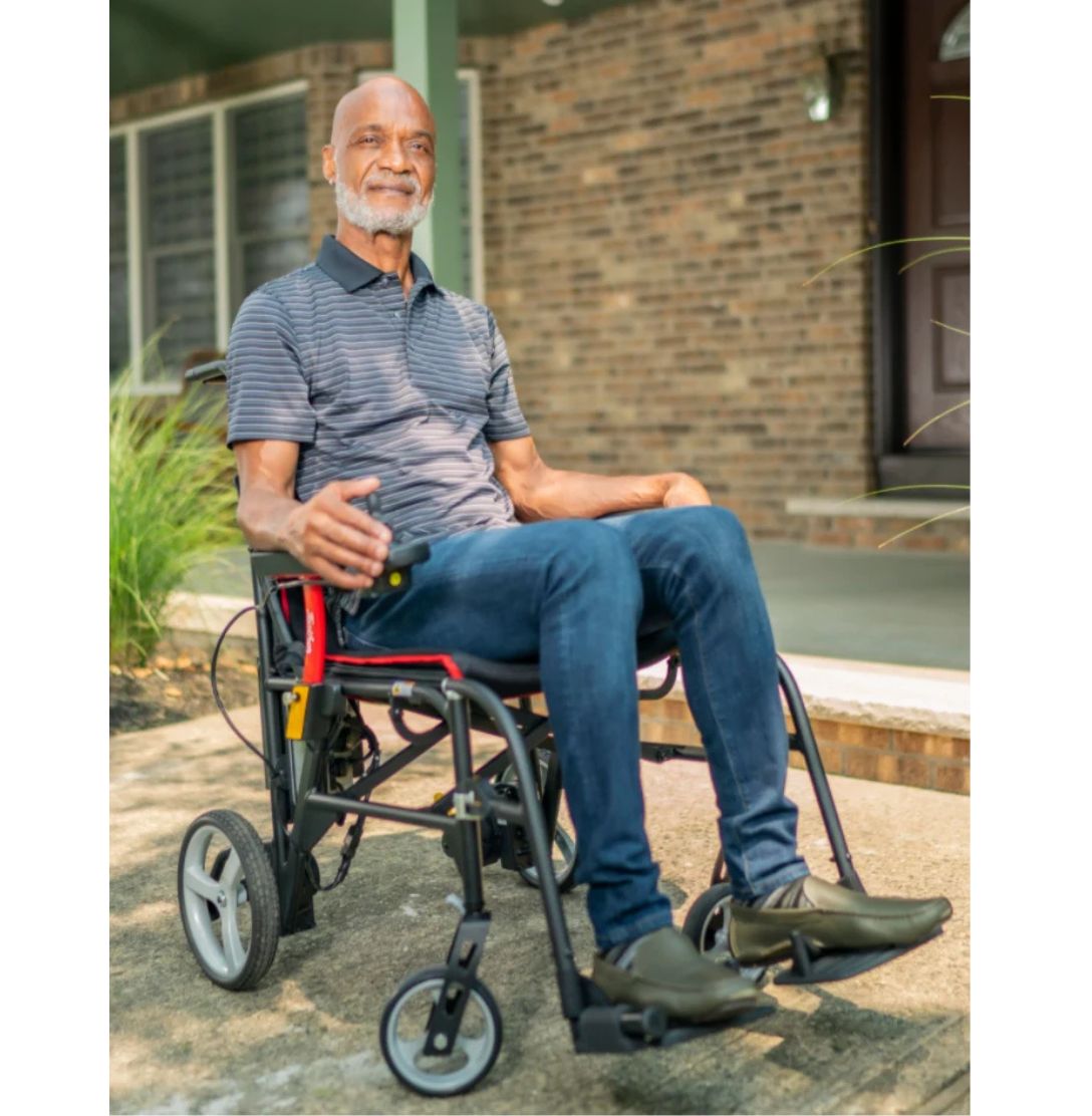 Man seated in a Feather Power Chair on a front walkway, hand on the joystick