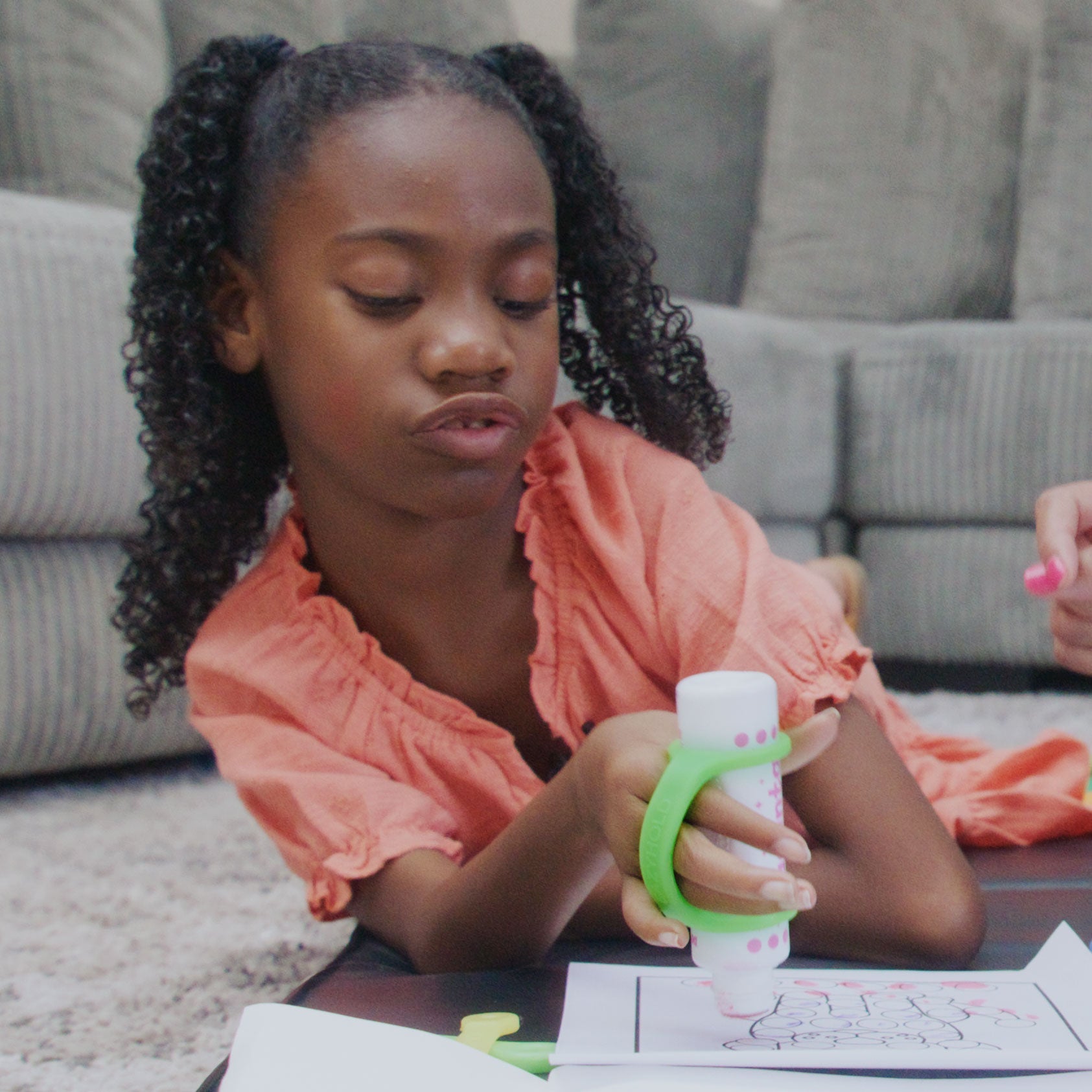 Young girl lying on the floor coloring with a marker held by a green EaZyHold silicone cuff