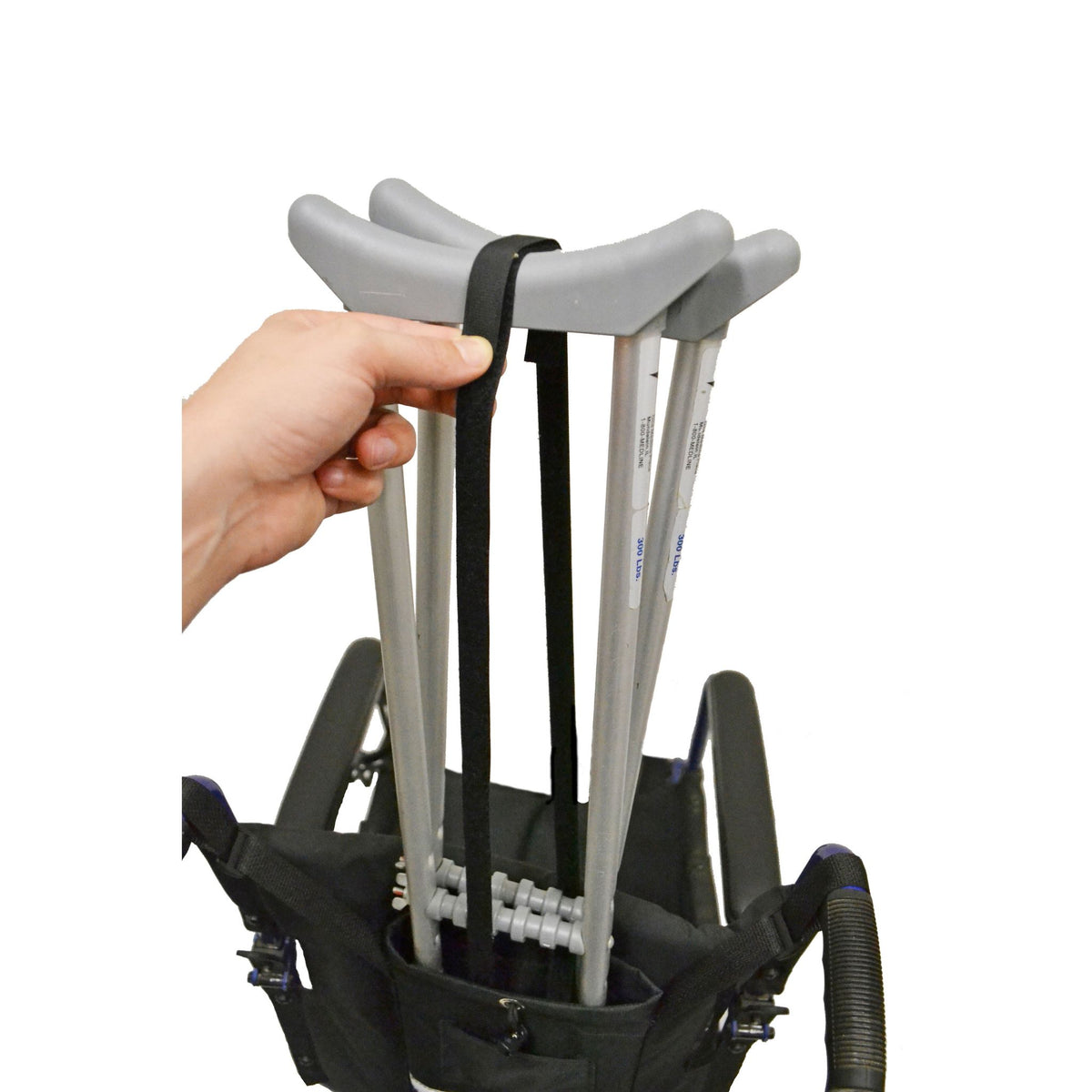 Person fastening the hook-and-loop strap around a pair of underarm crutches in the Diestco Crutch Holder behind a manual wheelchair seatback