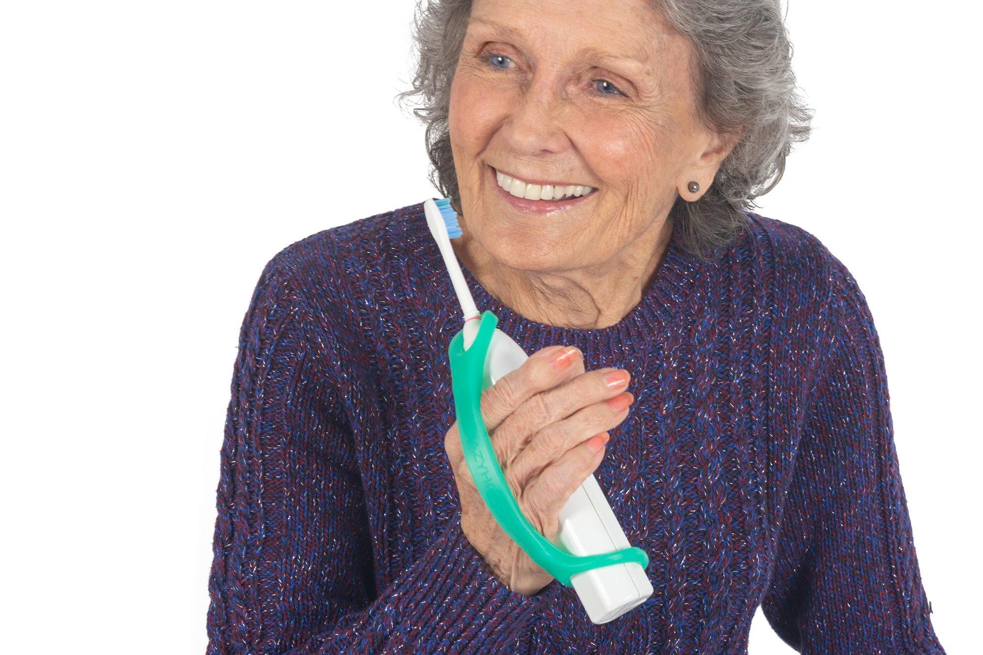 Smiling elderly woman holding an electric toothbrush with a teal EaZyHold silicone cuff wrapped around her hand