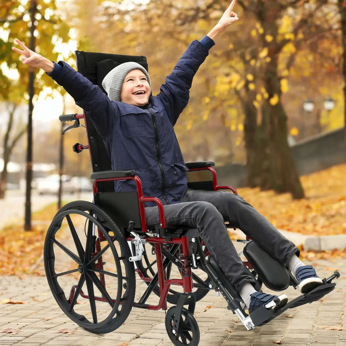 Happy boy with arms raised in a red Ziggo Pro reclining wheelchair with headrest and elevating legrests on an autumn park path