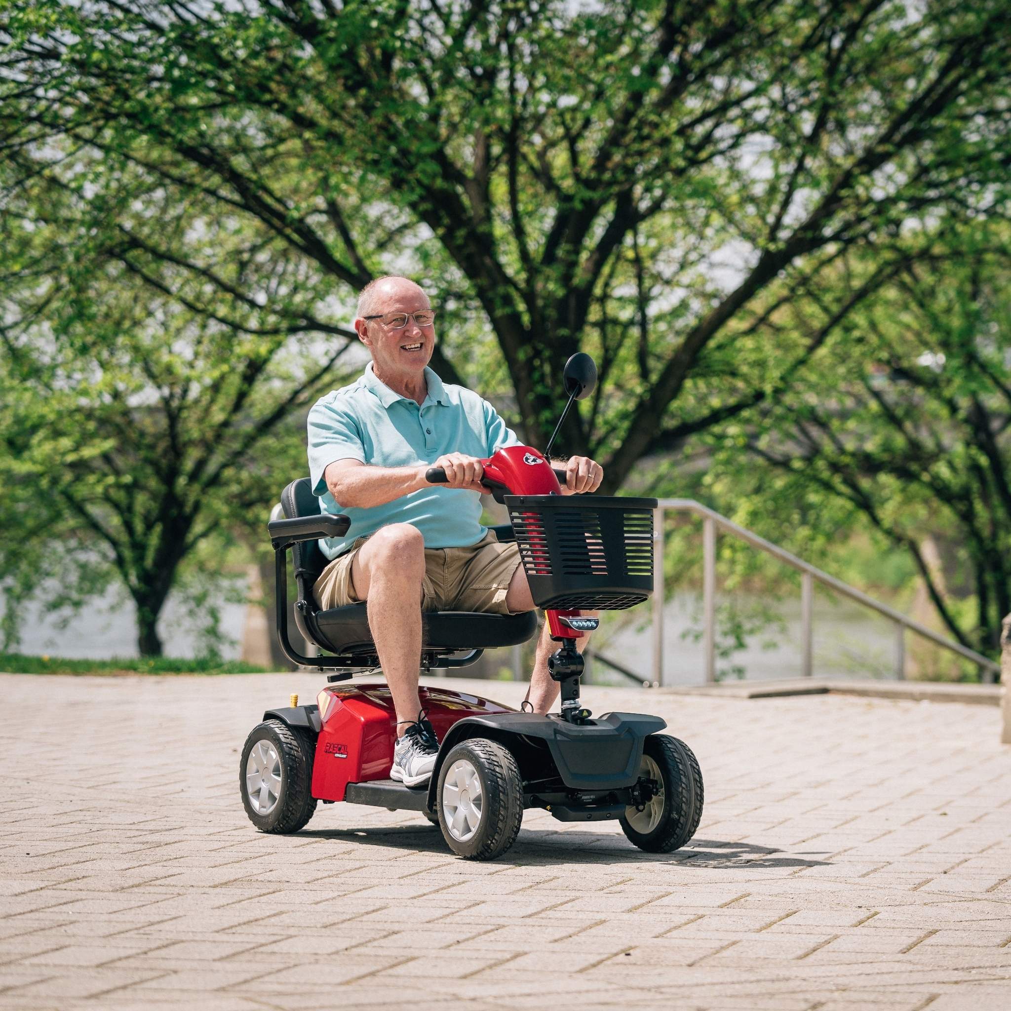 Adult riding the Rascal Conquest 4-wheel mobility scooter outdoors on a paved pathway, featuring a stable full-size design, comfortable captain’s seat, front basket, and everyday travel mobility performance.