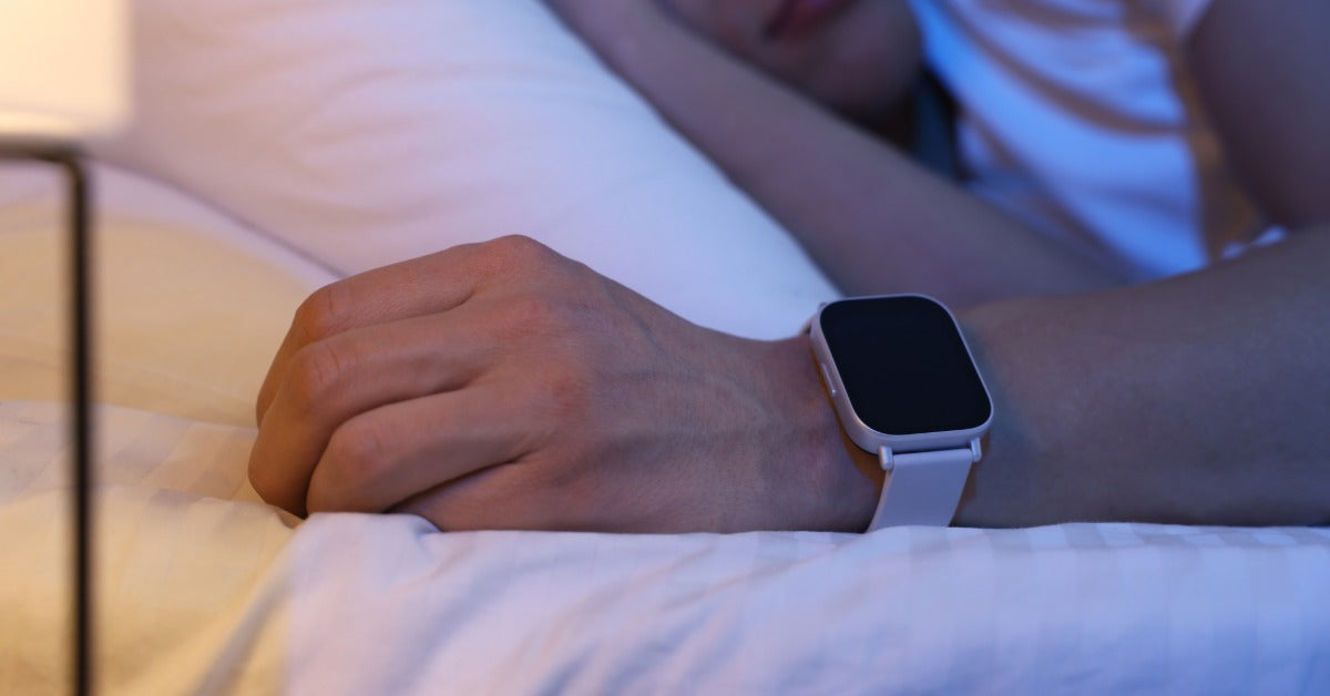 A person lying in a medical bed with white pillows and sheets. The person has a sleep tracker around their left wrist.