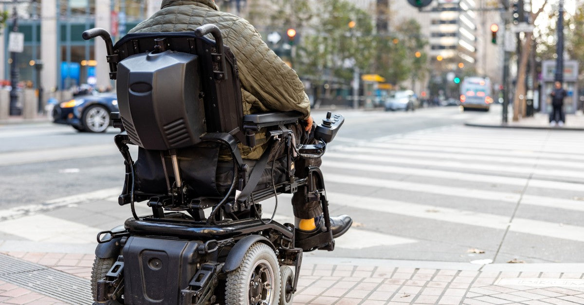 A person sitting in a wheelchair near a stoplight on the corner of a busy street during the daytime.