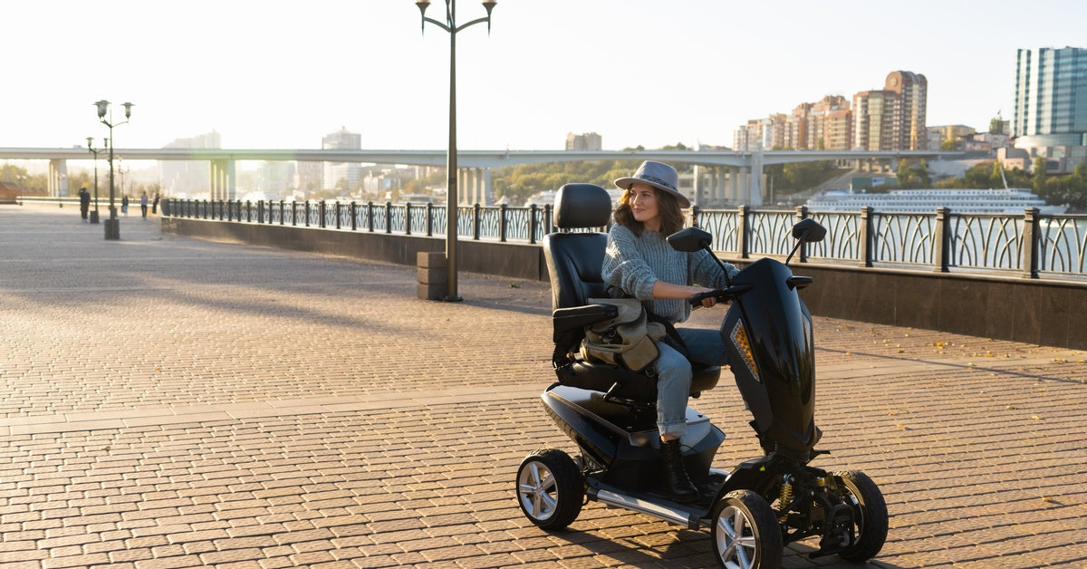 A person riding a black mobility scooter outside. The city skyline and a large bridge are visible behind them.