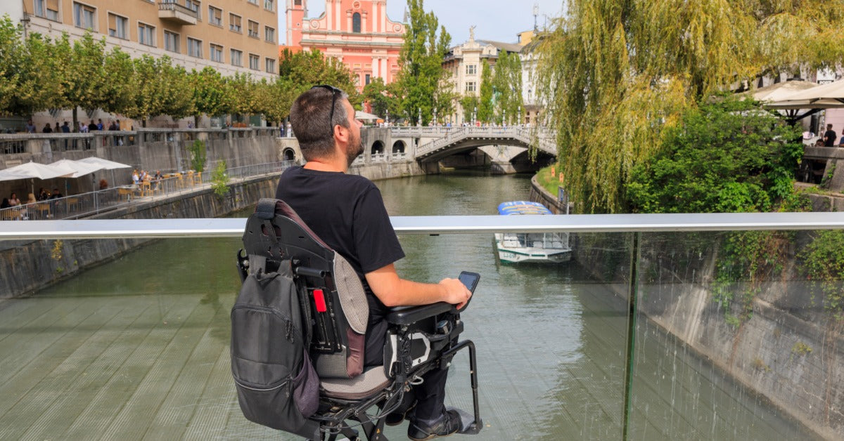 A person in an electric wheelchair outside on a bridge during the daytime. The bridge has metal railings.
