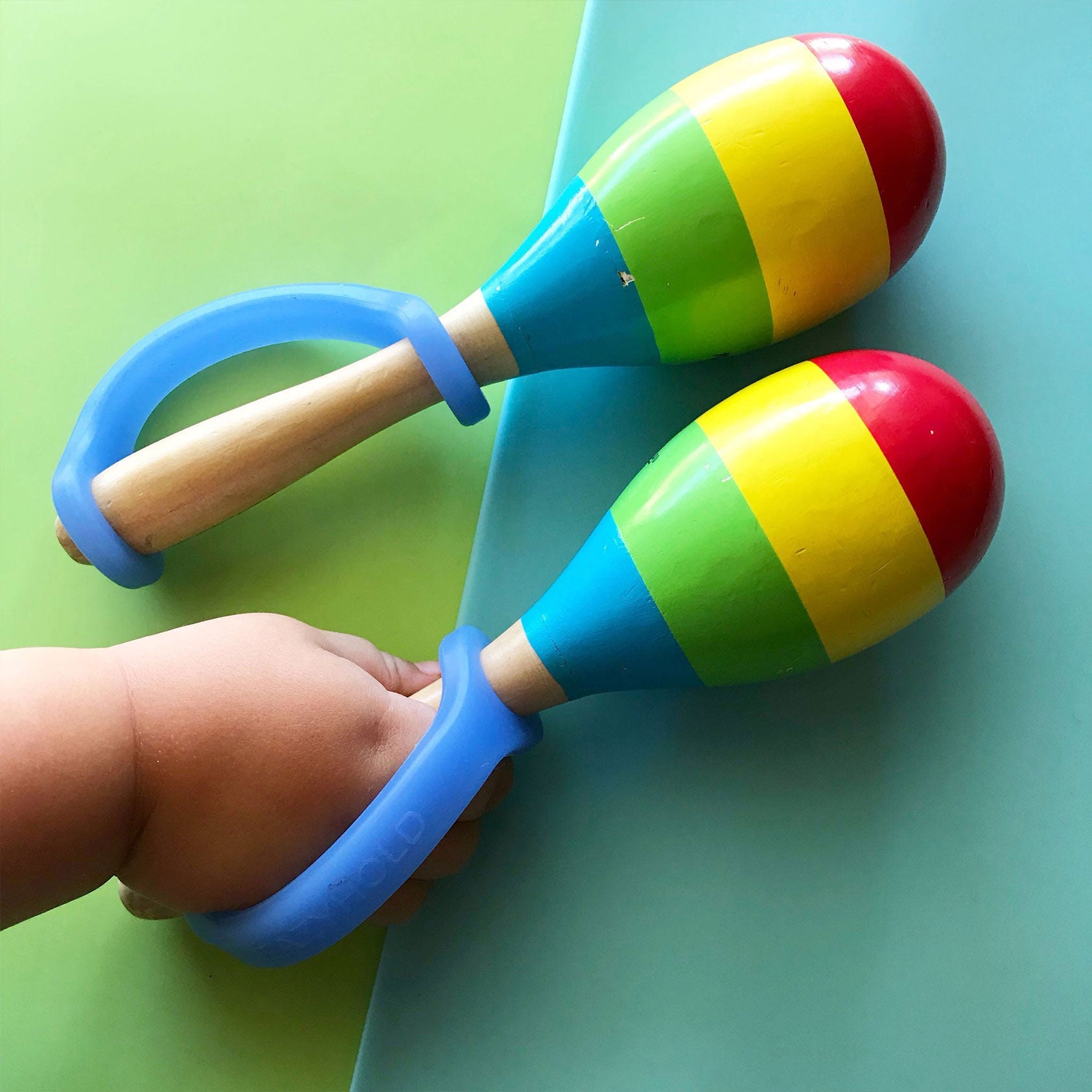 Infant gripping a wooden maraca with a blue EaZyHold cuff, with a second maraca alongside