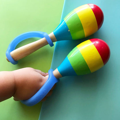 Infant gripping a wooden maraca with a blue EaZyHold cuff, with a second maraca alongside