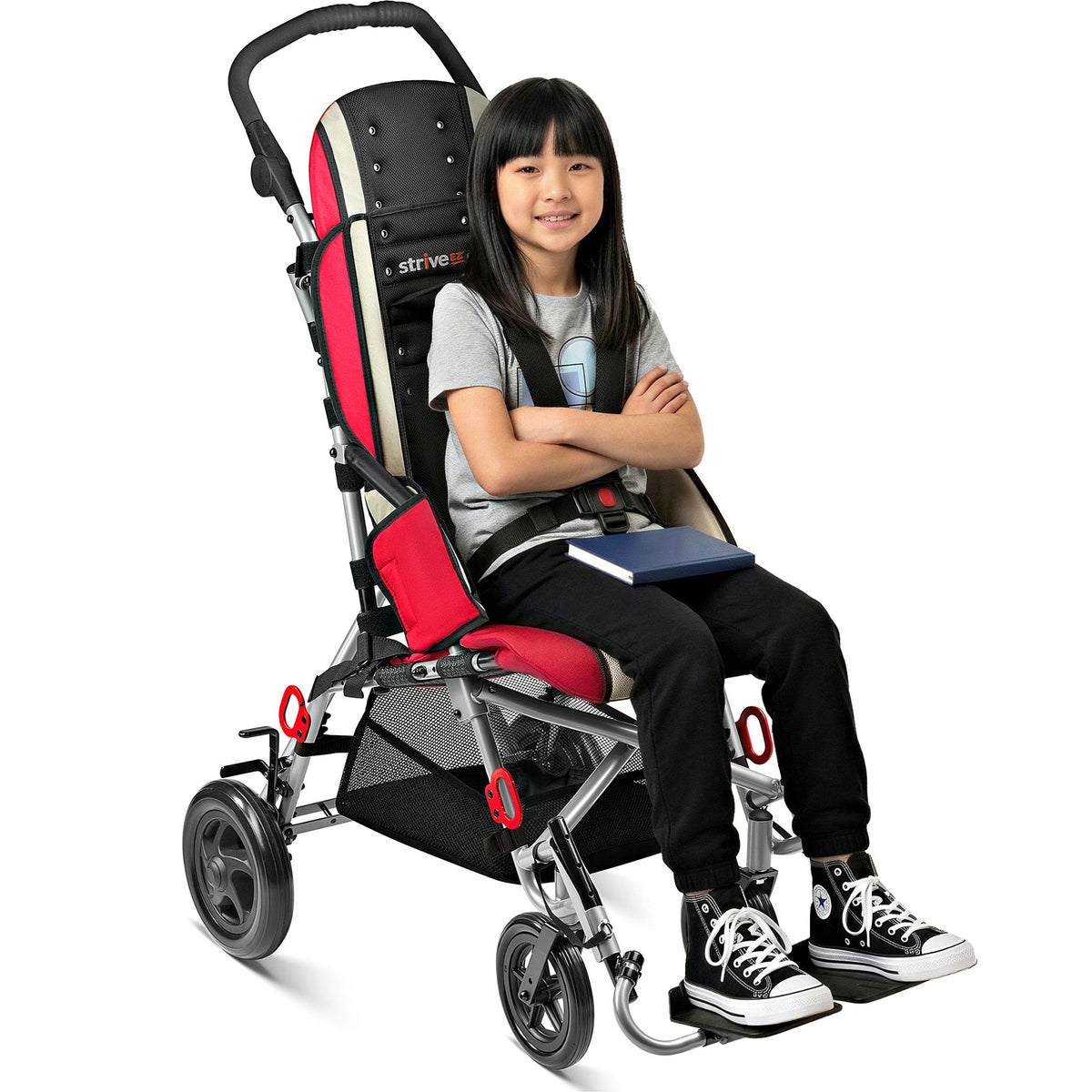 Smiling girl holding a book, seated in a red Circle Specialty Strive EZ adaptive stroller with 5-point harness and under-seat basket
