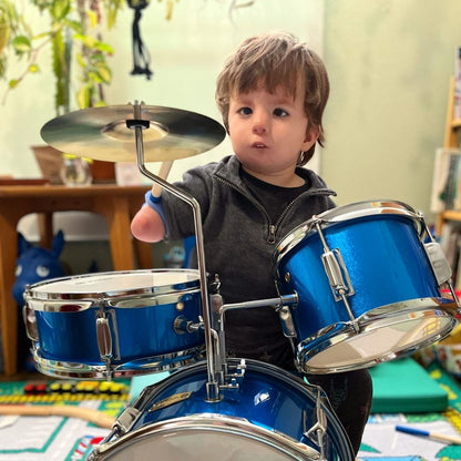Toddler playing a drum set, gripping a drumstick with a blue EaZyHold cuff