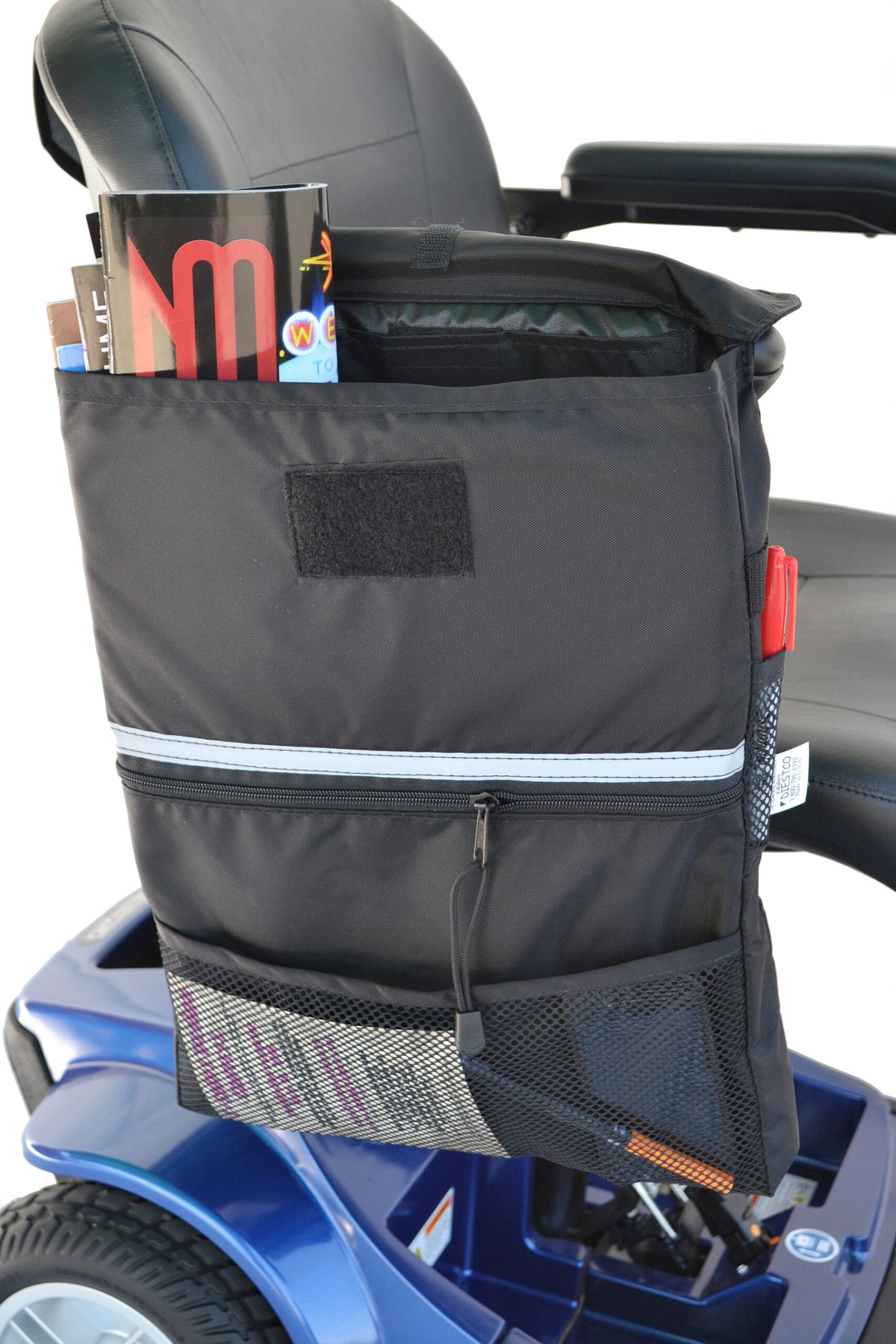 Diestco X-Large Saddle Bag hanging over a mobility scooter armrest, loaded with magazines in the top compartment, with a zippered pocket, mesh pockets, reflective strip, and hook-and-loop closure