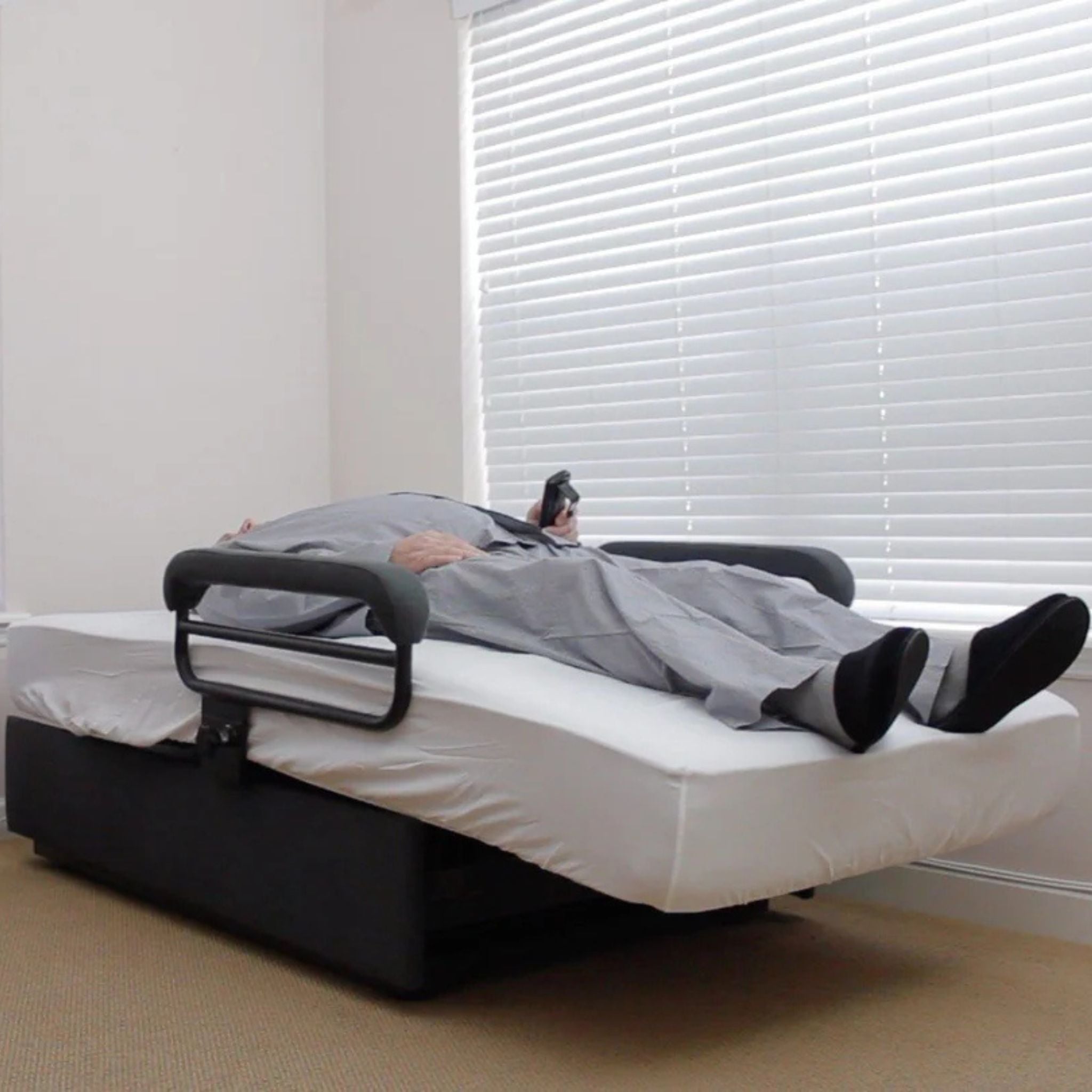 User resting in the ENVYY Sleep-to-Stand adjustable bed by Platinum Health in a flat sleeping position with supportive padded safety rails and remote-controlled positioning in a home bedroom environment.