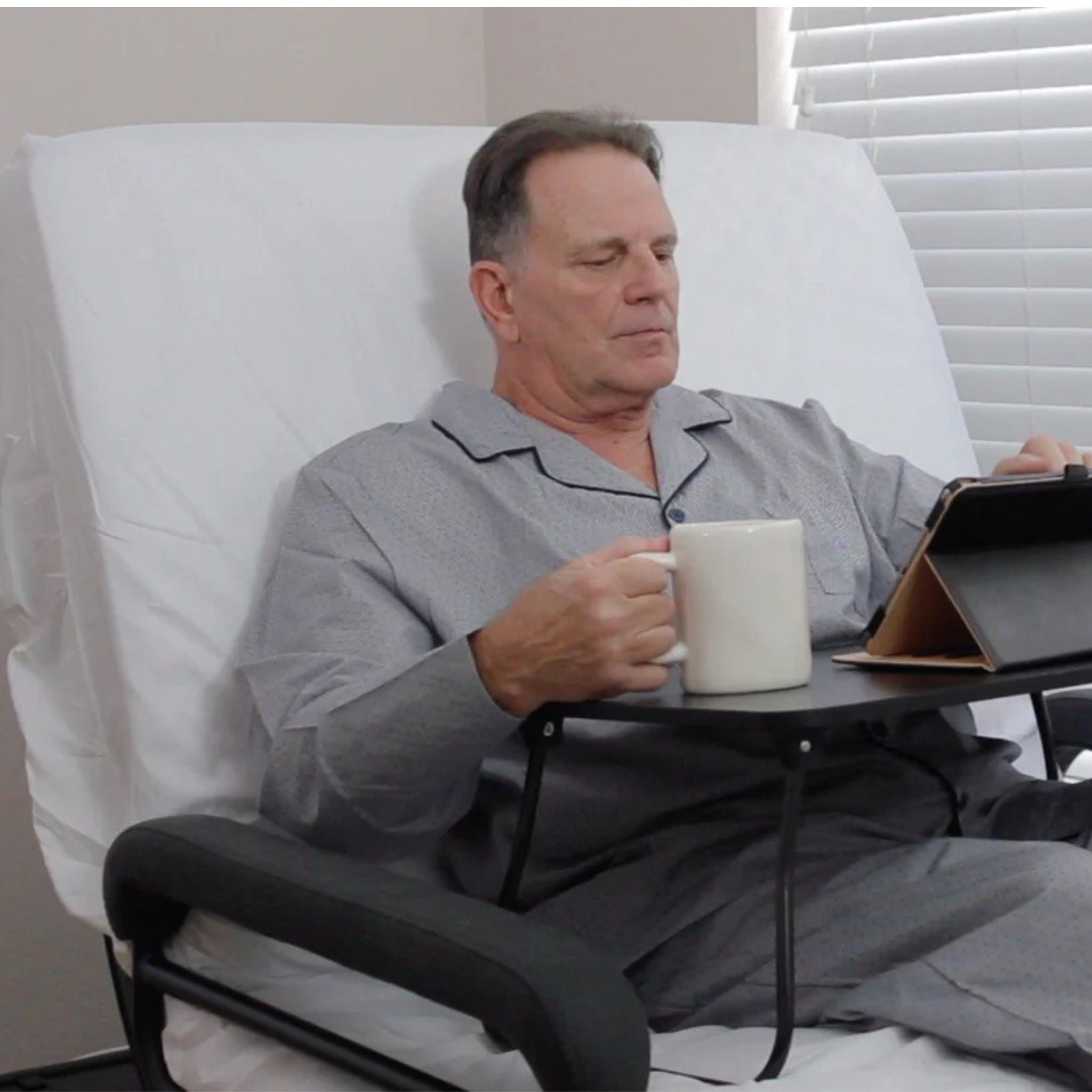 User sitting comfortably in the ENVYY Sleep-to-Stand adjustable bed by Platinum Health while drinking coffee and using a tablet, demonstrating supportive seated positioning for reading, meals, and everyday home activities.