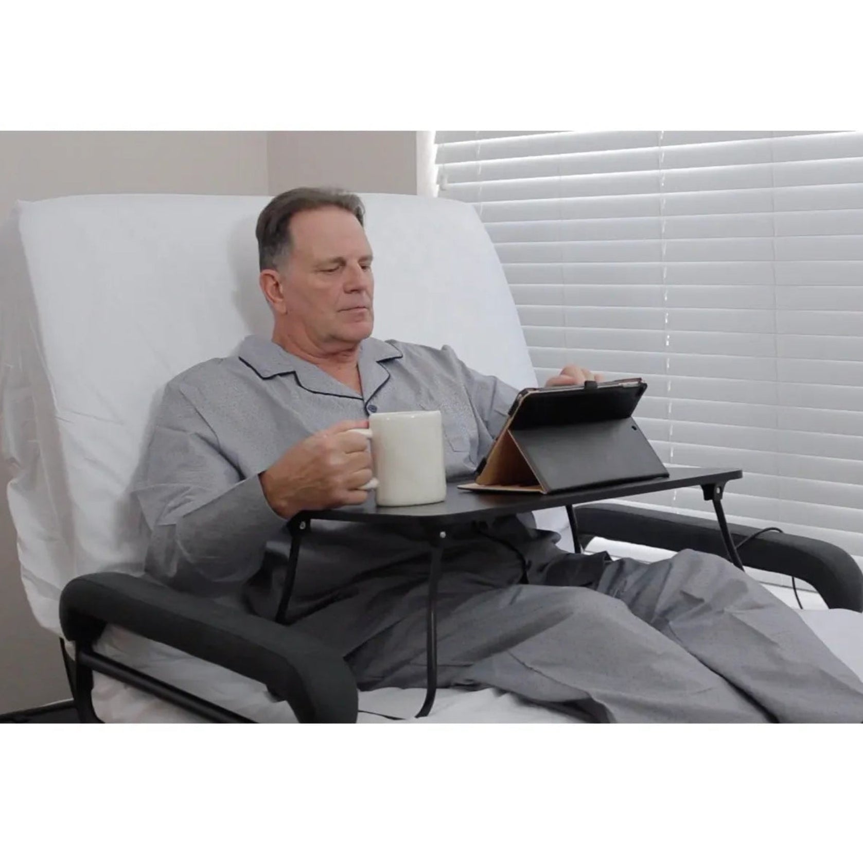 Man relaxing in an Envyy adjustable sleep-to-stand bed with padded side rails, using an overbed tray table with a tablet and coffee mug in a home bedroom setting.