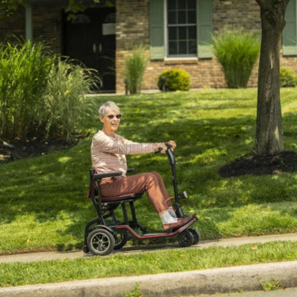 Woman riding the Feather Scooter along a sidewalk past a lawn and front garden