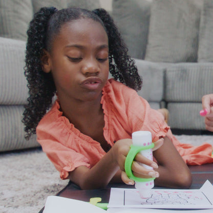 Young girl lying on the floor coloring with a marker held by a green EaZyHold silicone cuff