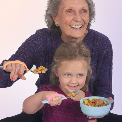 Smiling older woman and young girl each using EaZyHold cuffs on spoons to eat cereal independently