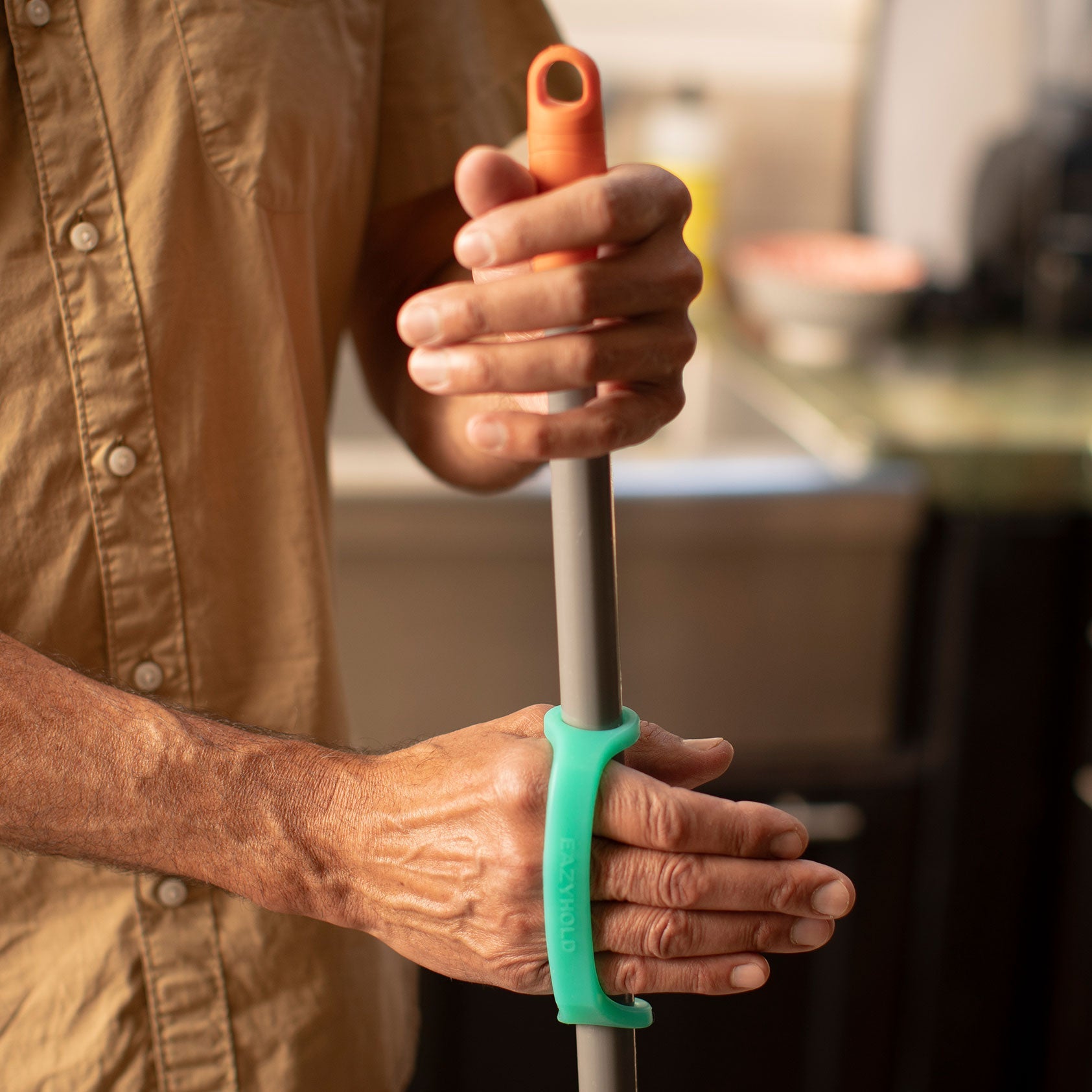 Man in a kitchen using aqua and teal EaZyHold cuffs to grip a long broom or mop handle with both hands