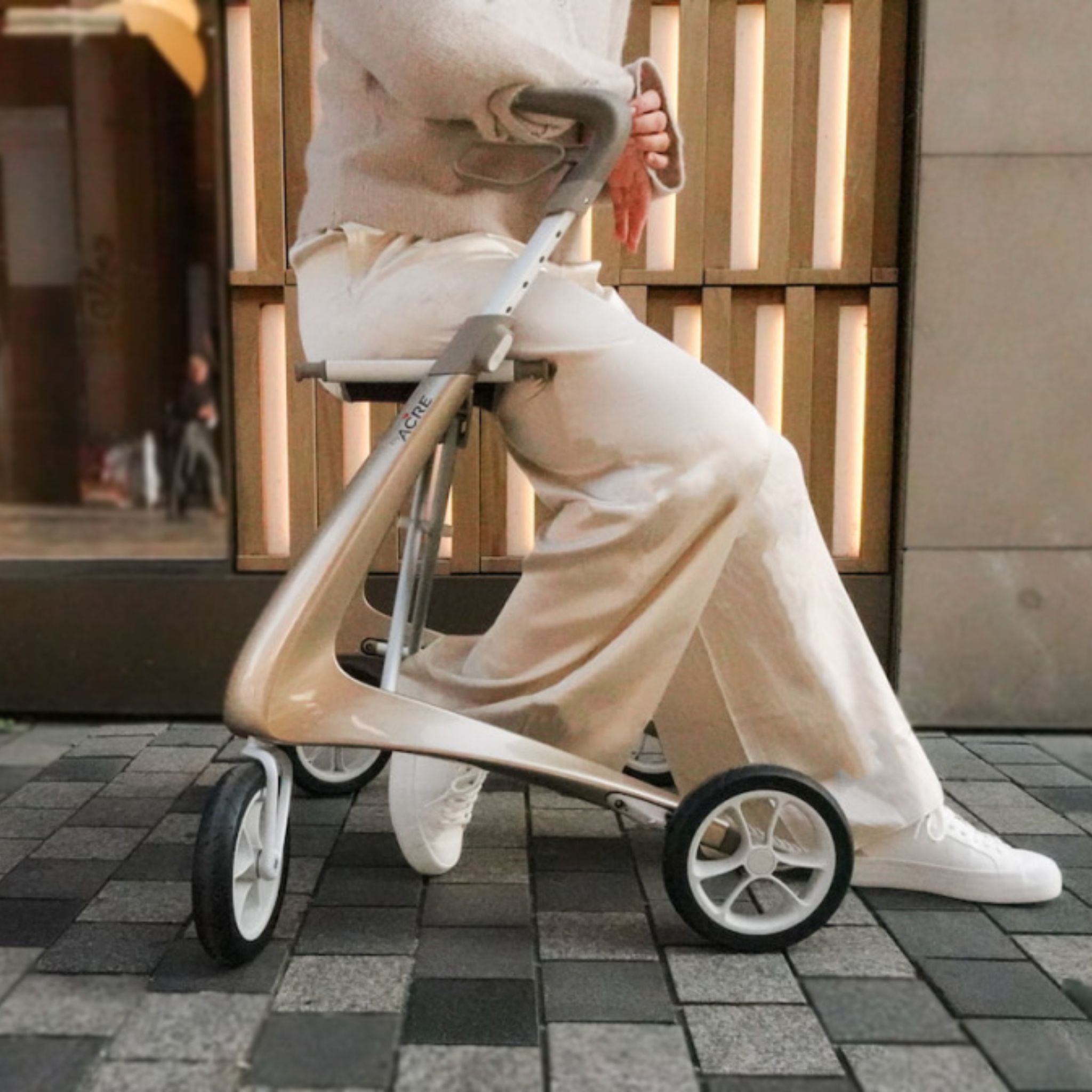 Person walking with a Champagne Gold byACRE Carbon Ultralight rollator along a city sidewalk