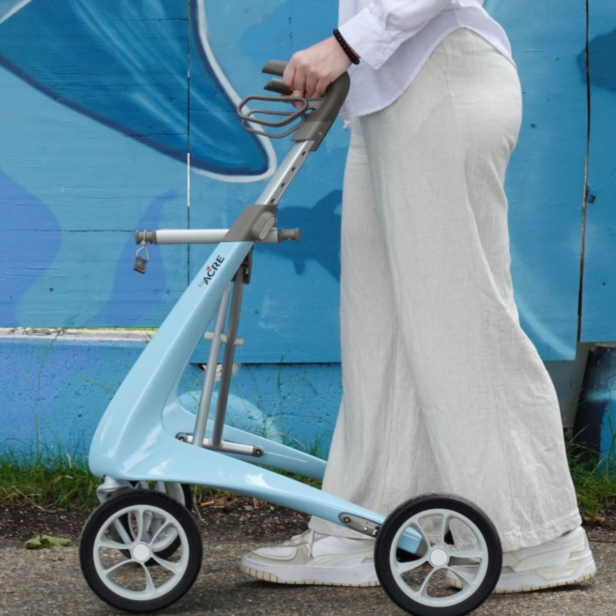 Person walking with a Morning Blue byACRE Carbon Ultralight rollator past a blue painted mural wall