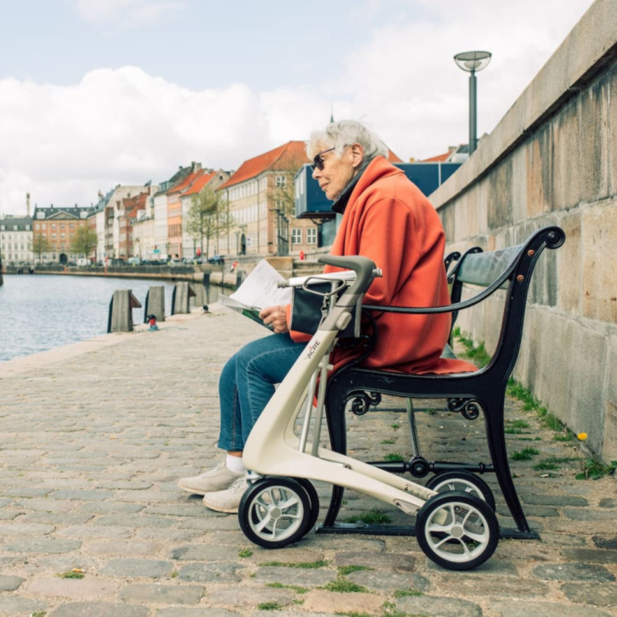Woman resting on a bench beside an Oyster White byACRE Carbon Ultralight rollator by the water