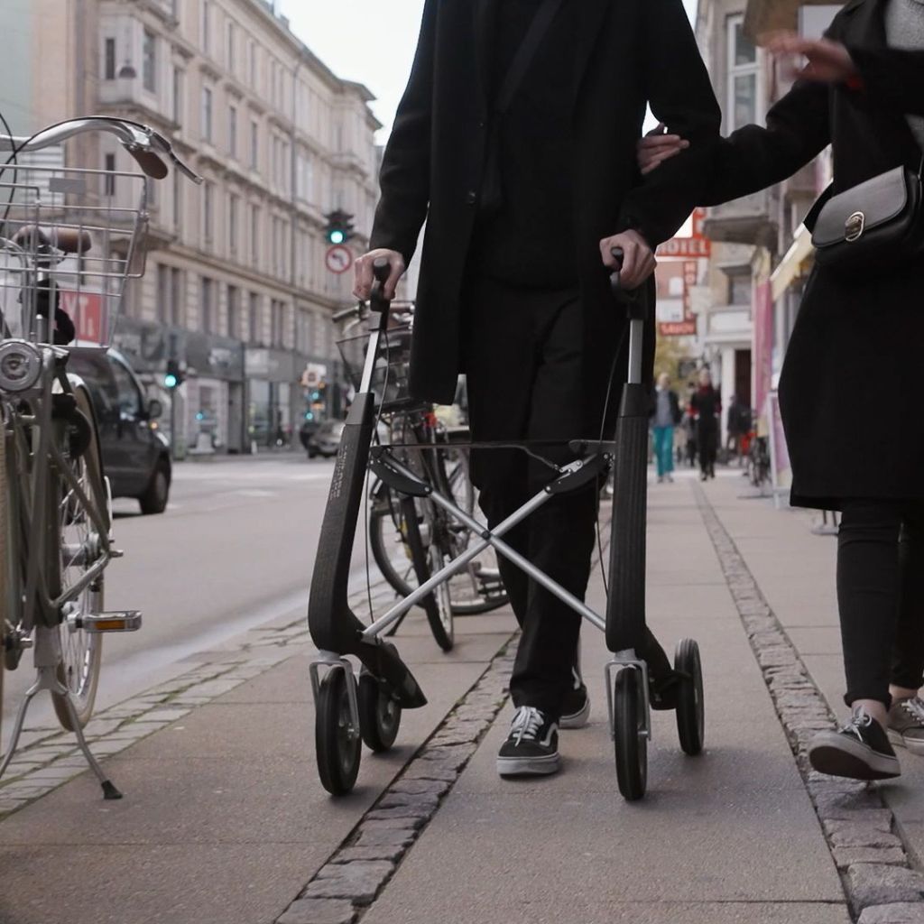 Person walking with a Granite Black byACRE Nordic Pioneer rollator along a city sidewalk