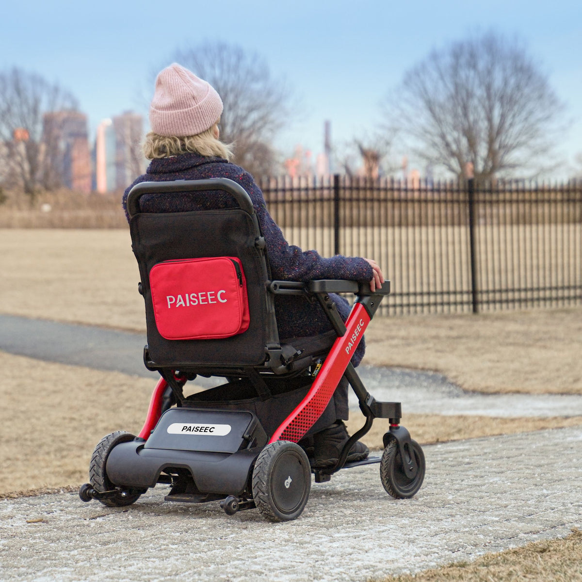 Woman riding a red Paiseec W1 electric wheelchair along a park path with city skyline in the background